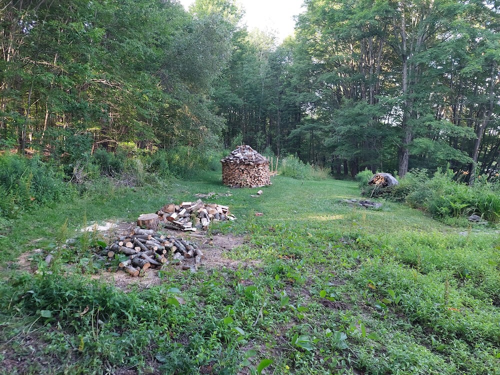 Firewood processing area showing a pile of small tree branch rounds yet to be split, a chopping block, and some split firewood. A rotund holz hausen, or wood house, is featured in the background and represents all the wood from the larger maple logs.