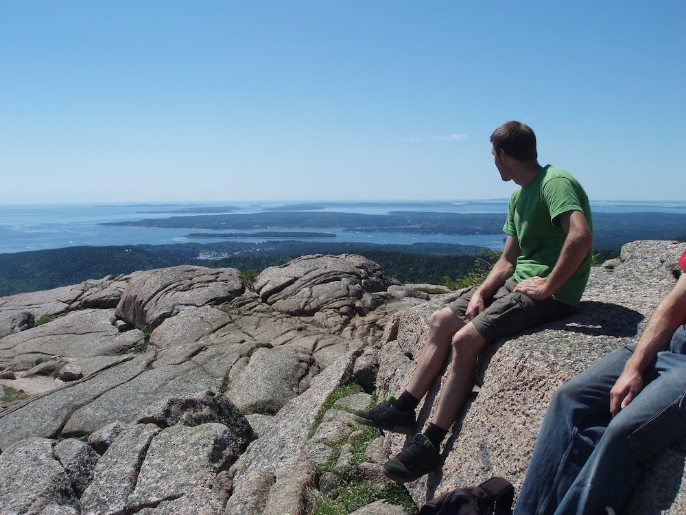 A man on top of an unvegetated solid rock mountain top looking at ocean and land in the distance.