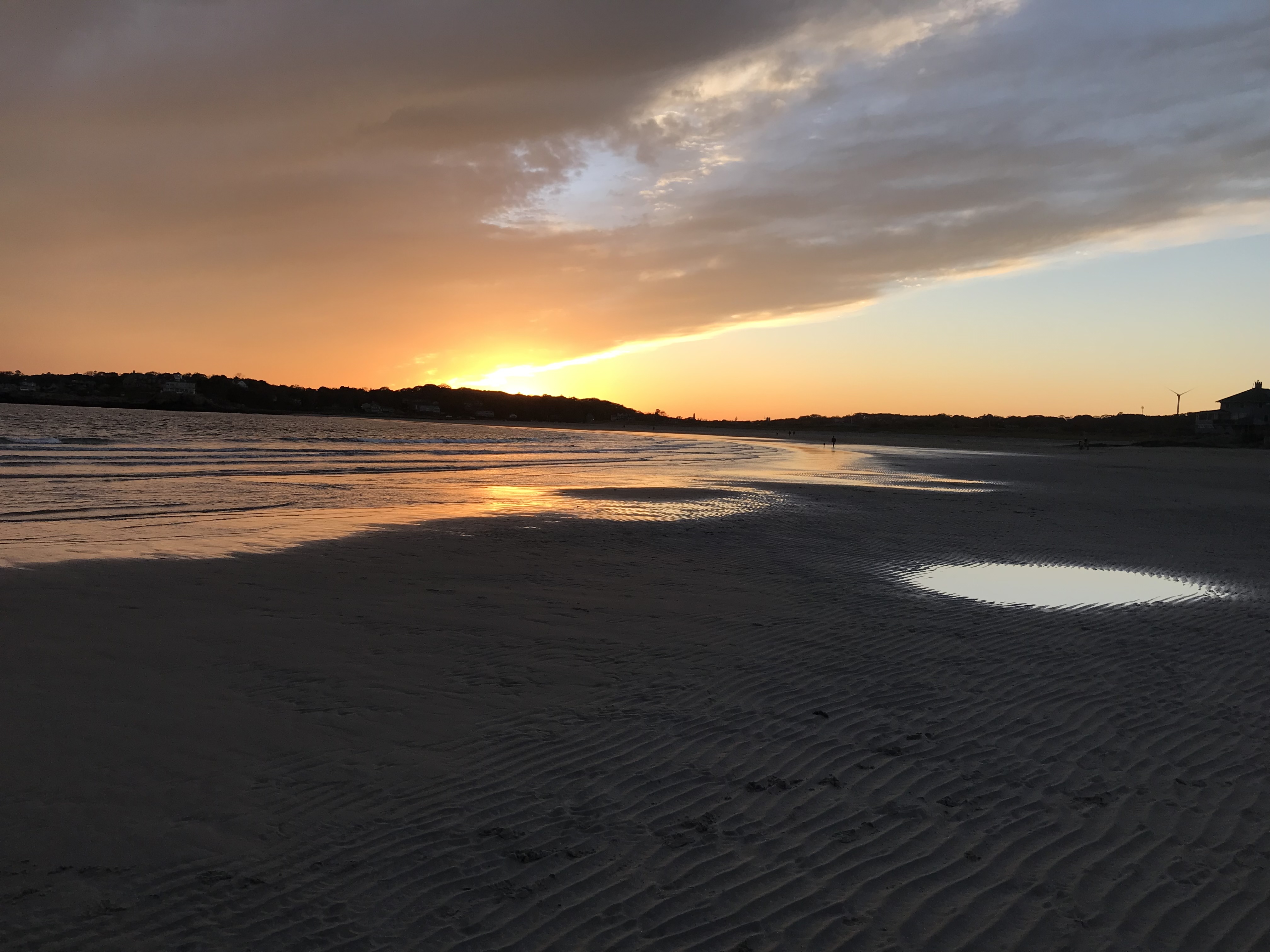 The ocean in a bay with a sandy shore, set ablaze with orange color from a brilliant sunset over the nearby hill with a mostly cloudy sky, colored different shades of orange and yellow.