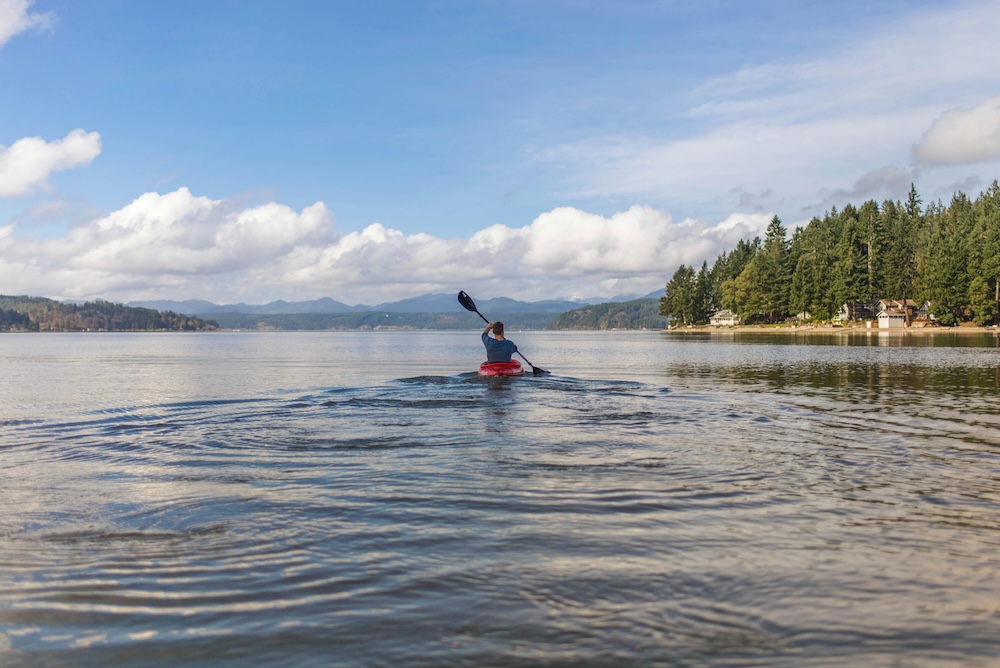 Man paddling a red kayak in a large lake with cottages on the wooded shoreline, blue sky with puffy white clouds and mountains in the background.  