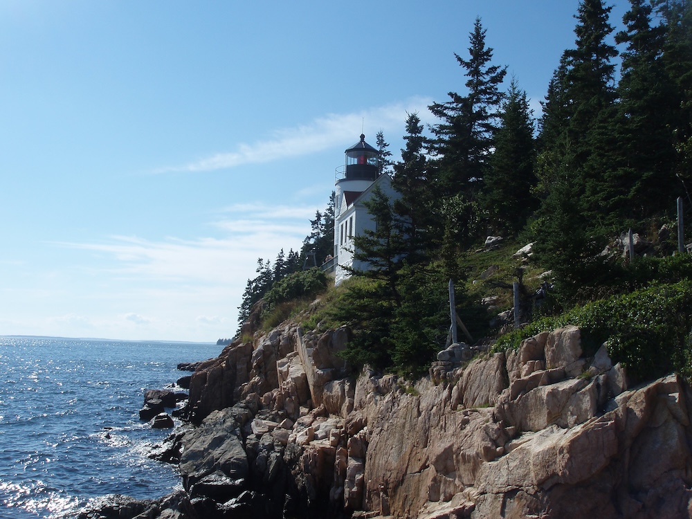 Lighthouse on the edge of a solid rock ocean shoreline with conifer trees around it.