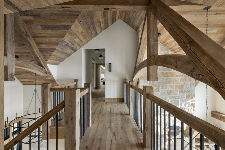 A second-floor catwalk with exposed curved beams and rafters and a beautiful blend of different shades of wood ceiling boards, wood posts and rails. A wood floor is offset with white walls.