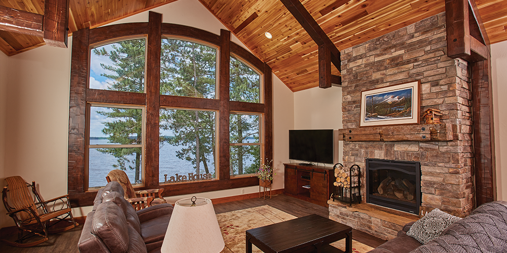 Arched living room area with large stone fireplace, wide wood-framed window, wooden rafters and wood ceiling and floor.