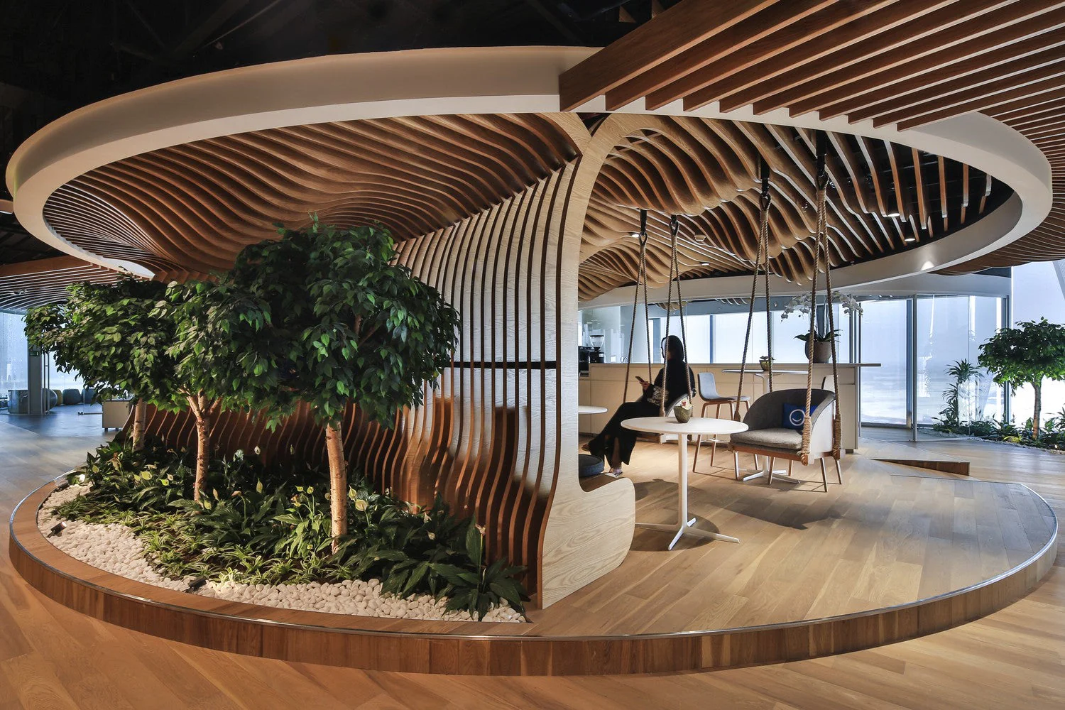 Raised circle area with plants, tables and swings for chairs. Undulating wood slats in the ceiling and a wall. The floor is also made of wood.