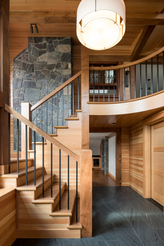 A beautiful wooden staircase with a landing and wide stone chimney.  View down a hall with stone floor and wood trimmed walls.