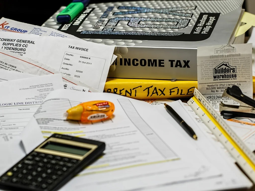 A messy desk covered with income tax return books, paperwork, and office supplies. 