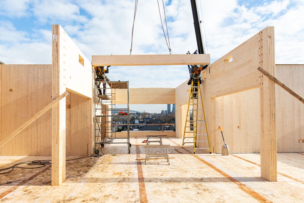 Mass timber panels in place to create walls with a beam being held by a crane and two men putting it in place in a hallway.