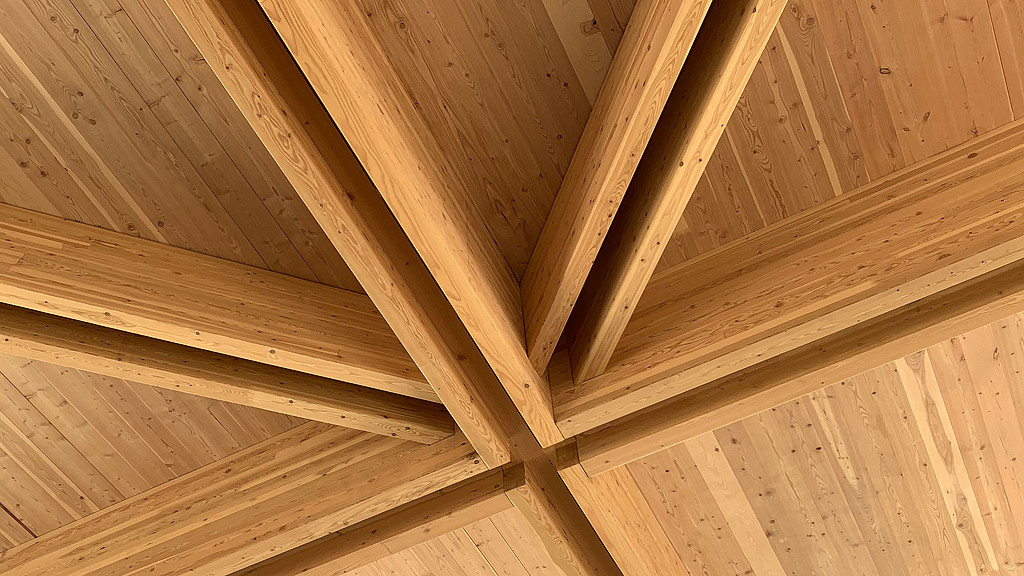 View of beautiful mass timber supporting beams joining at different angles and supporting an exposed wood panel ceiling.  