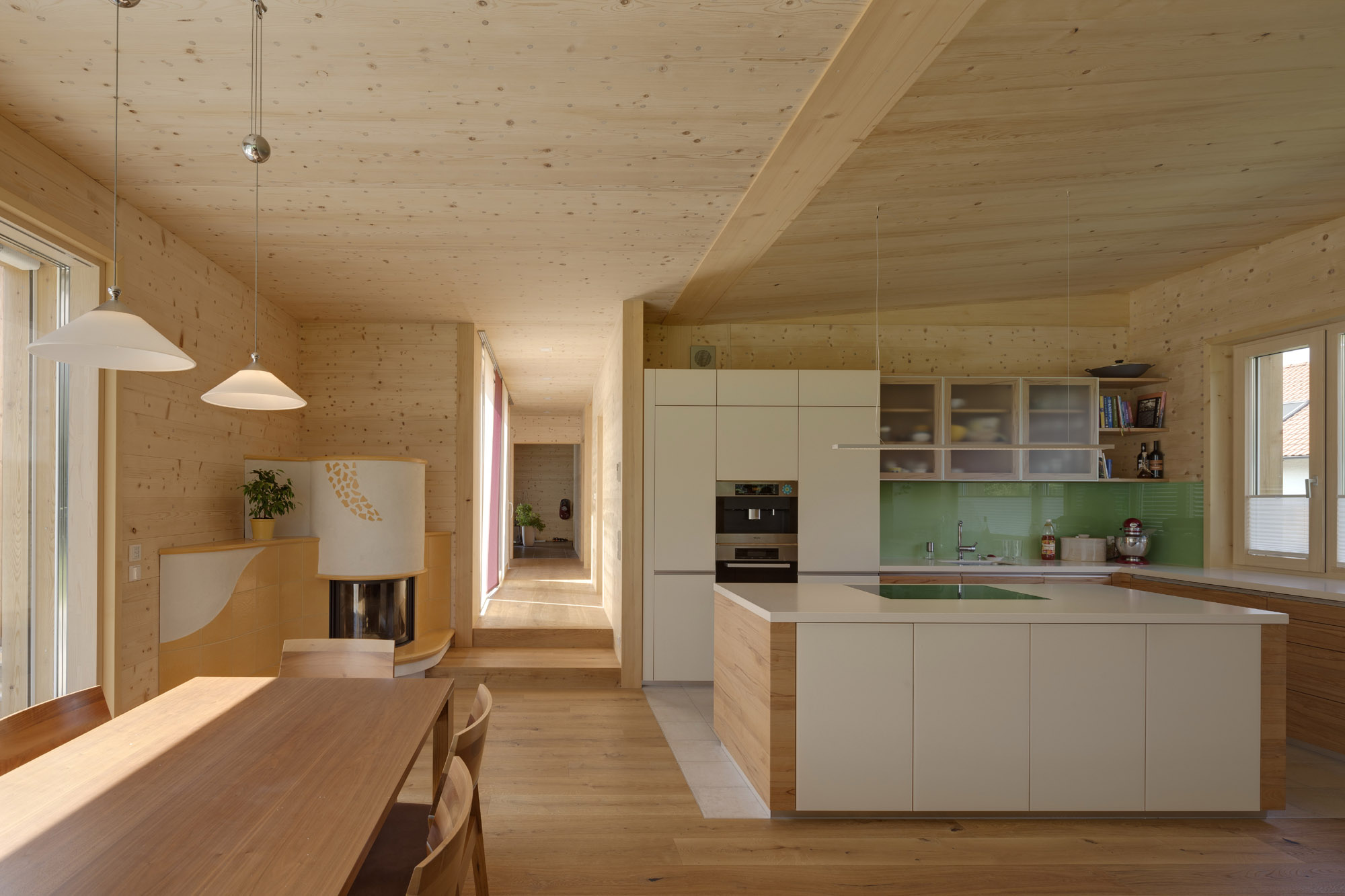 View of a kitchen dining area with much exposed wood on the floor, walls and ceiling and a wooden table and chairs.  There is a kitchen island and view down a long hallway.