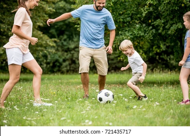 A mom and dad kicking a soccer ball with their two children with the woods in the background.