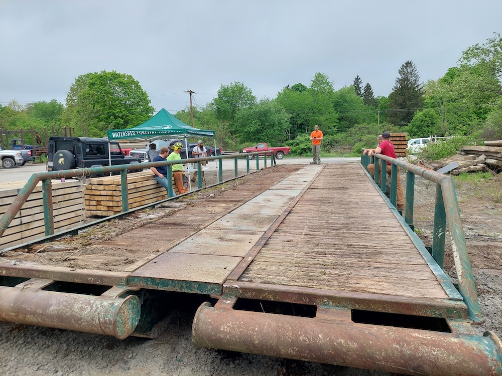 A 50-foot-long, hinged portable bridge with rotten decking. The new decking (made of red oak) is stacked beside the bridge. Some workshop participants are talking while they wait for the workshop to begin.