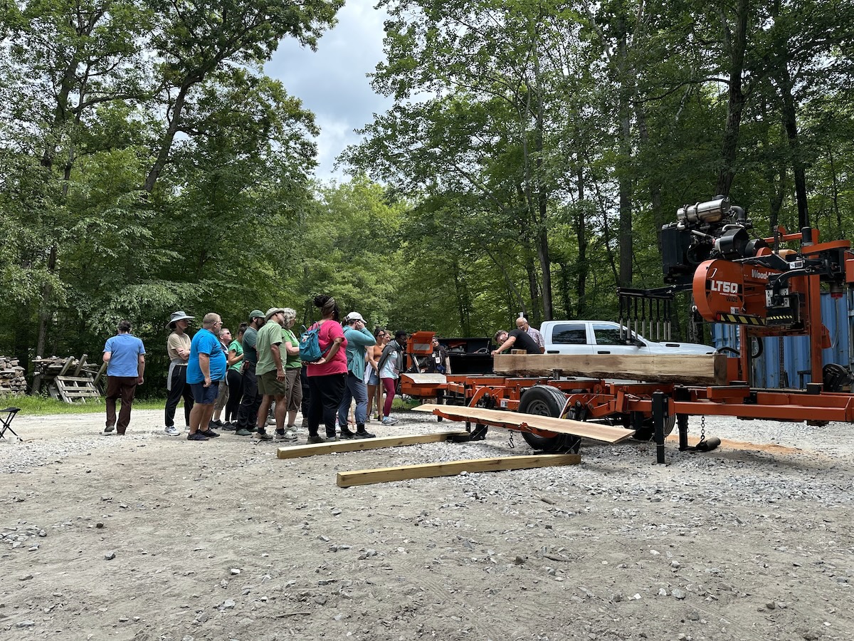 A group of people watch someone mark the thickness of boards to be cut from a cant placed on a portable sawmill. 