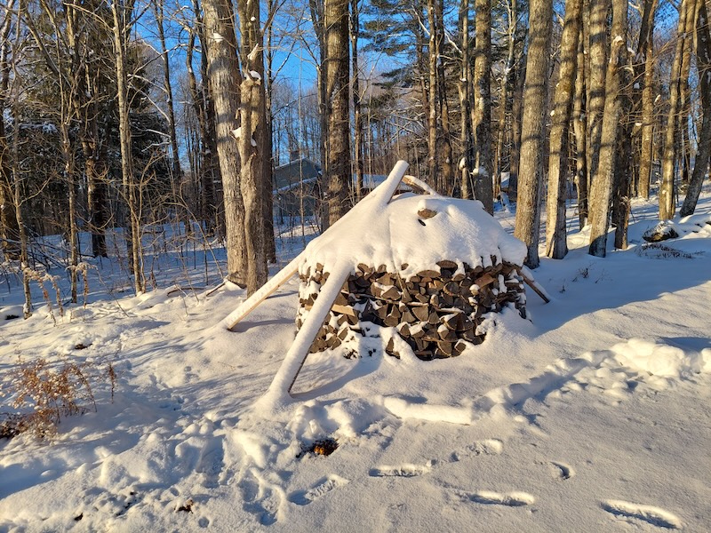 A beehive-shaped stack of firewood covered in snow and bathing in morning sunlight.