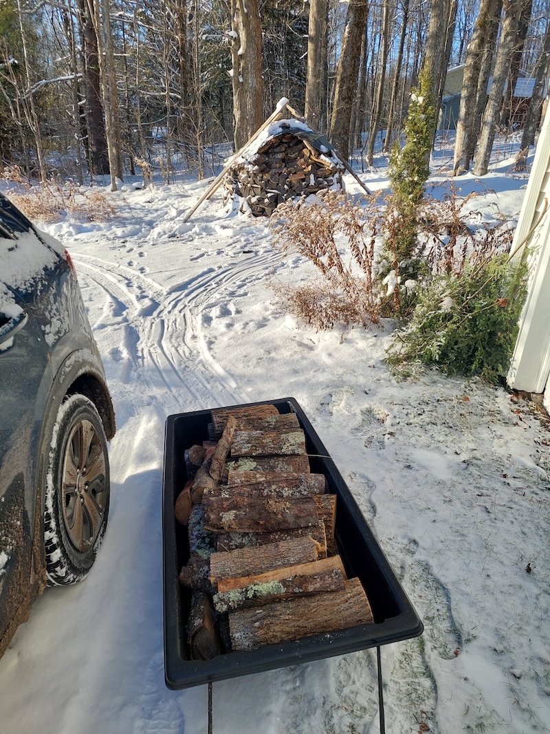 A jet sled full of firewood getting pulled over the snow-covered driveway to the breezeway. 