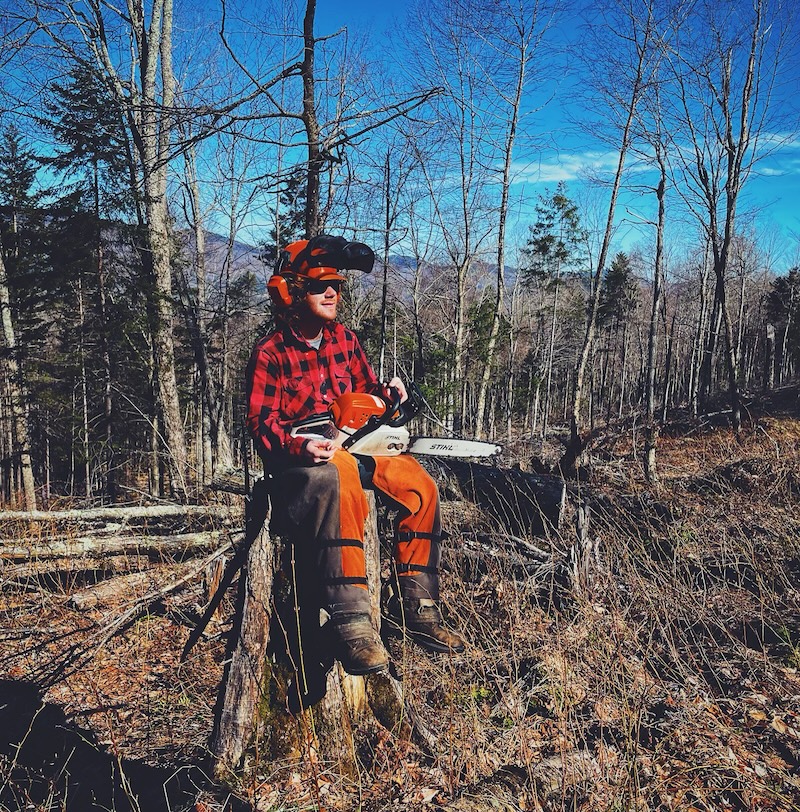 Image showing a man sitting on a tree stump holding a chainsaw in an area with felled trees wearing a hard hat, glasses, ear protection, chainsaw chaps.